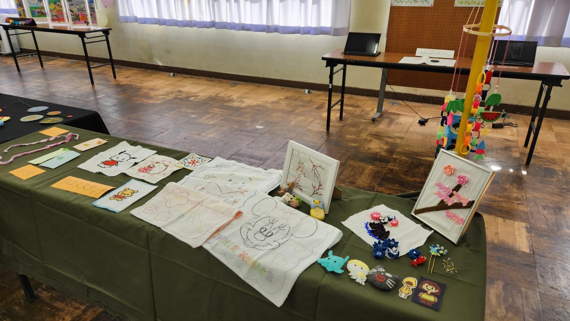 ハンドクラフト部が作成したディズニーのキャラクターなどを描いたクロスステッチ刺繡やミサンガなどが濃い緑色のテーブルクロスがかけられた長机の上に展示されている作品の写真