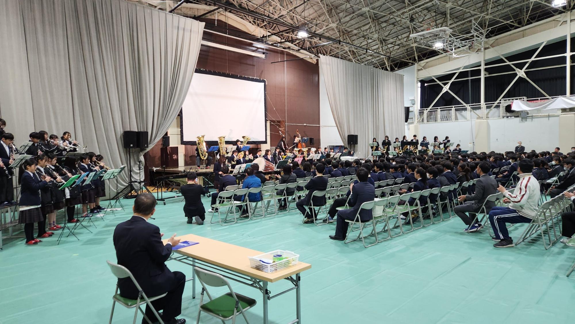 体育館のステージや左右に広がっている吹奏楽部の演奏を、生徒や教師がパイプ椅子に座って聞き入っている様子の写真