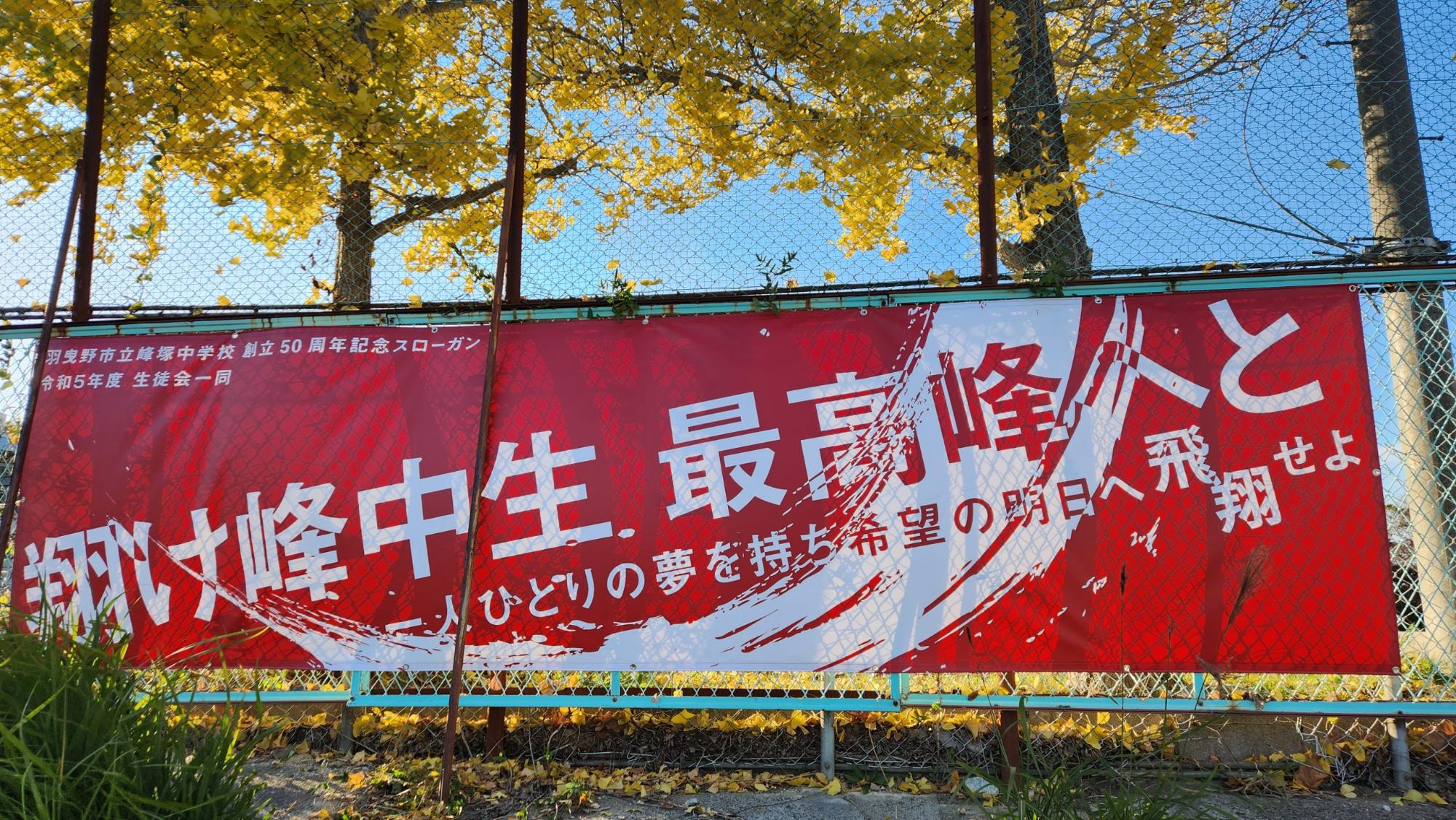 フェンスに設置された赤地に白文字で「翔け峰中生、最高峰へと 一人ひとりの夢を持ち希望の明日へ飛翔せよ 羽曳野市立峰塚中学校 創立50周年記念スローガン 令和5年度 生徒会一同」と書かれた大きな横断幕の写真