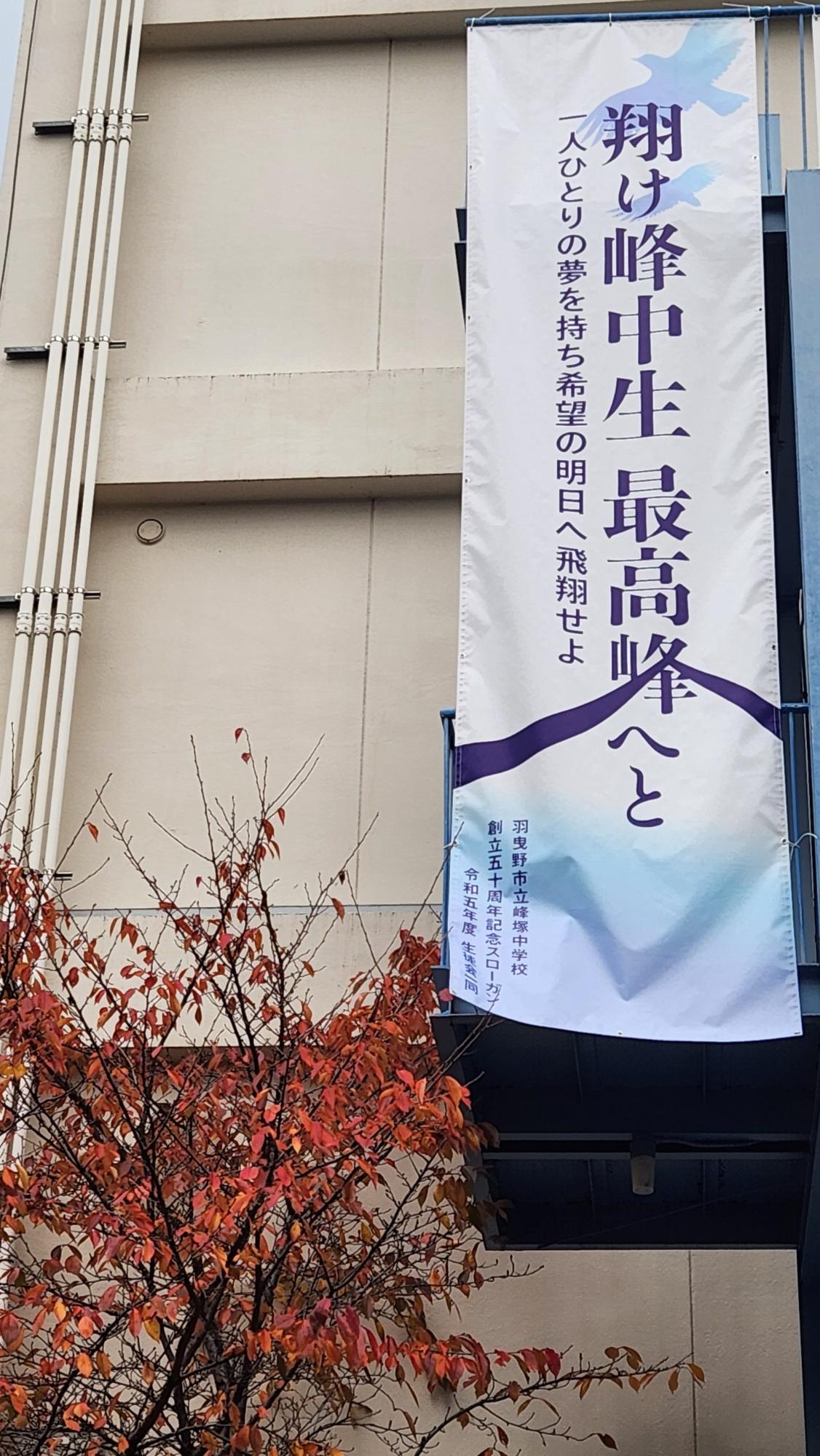 白地に紫文字で「翔け峰中生、最高峰へと 一人ひとりの夢を持ち希望の明日へ飛翔せよ 羽曳野市立峰塚中学校 創立五十周年記念スローガン　令和五年度　生徒会一同」と書かれた大きな懸垂幕が校舎外側に設置されている写真