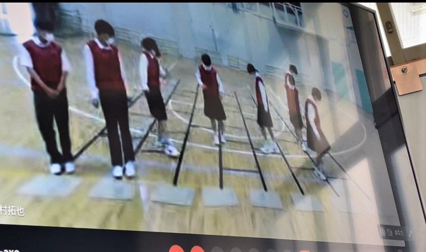 体育館の床にできたあみだくじの線の上に赤色のビブスを着た7名の生徒が立っているクラスカラー決めの様子の写真