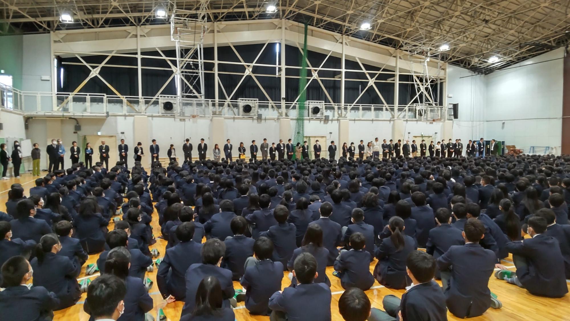 体育館の前方壁側に教職員が一列に並んで立ち、床に座った生徒たちが話を聞いている教職員紹介の様子の写真