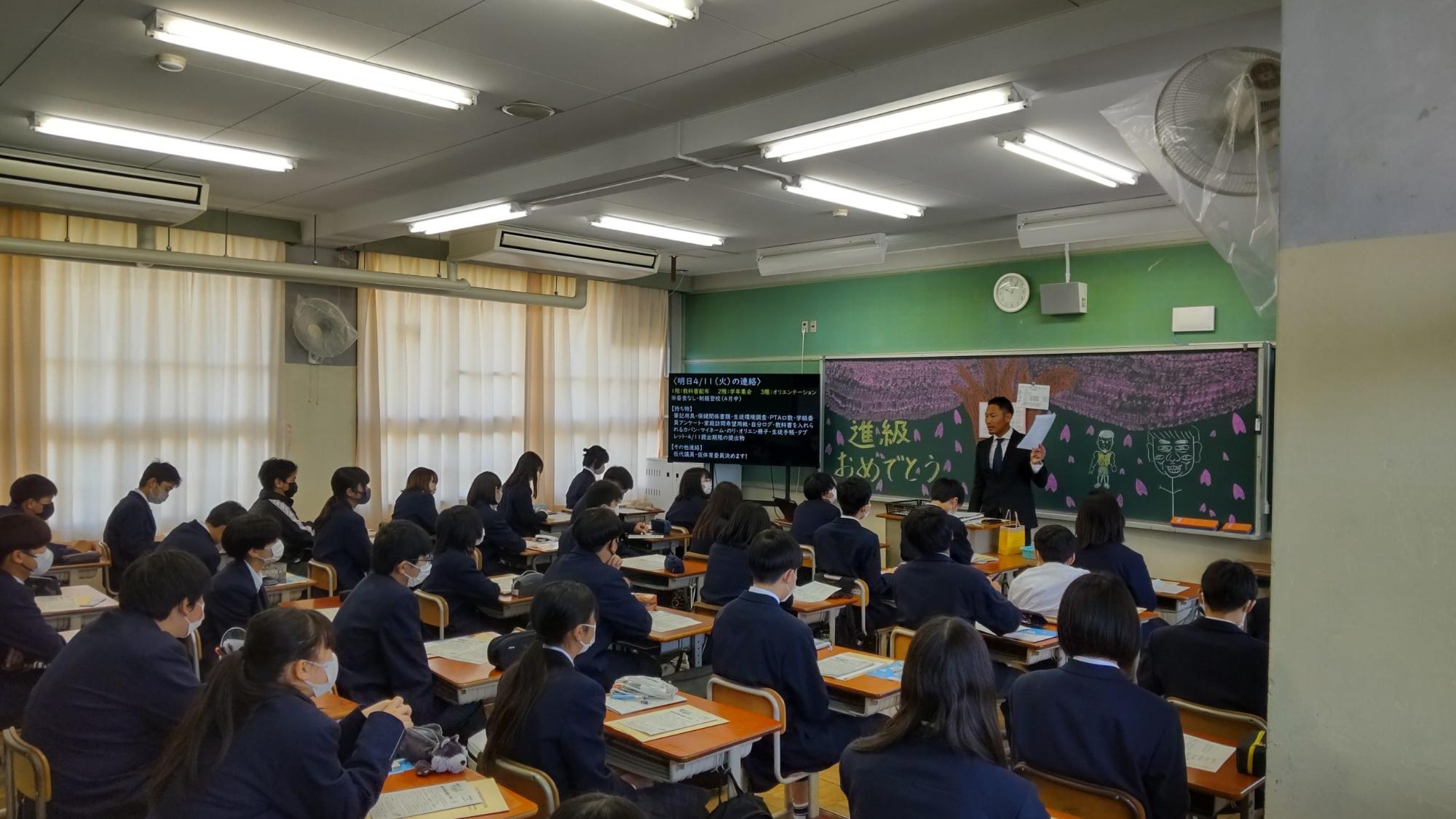 黒板いっぱいに桜の木と花びら「進級おめでとう」とチョークで書かれた教壇に男性教師が立ち、自席に座った生徒たちが話を聞いている教室の様子の写真