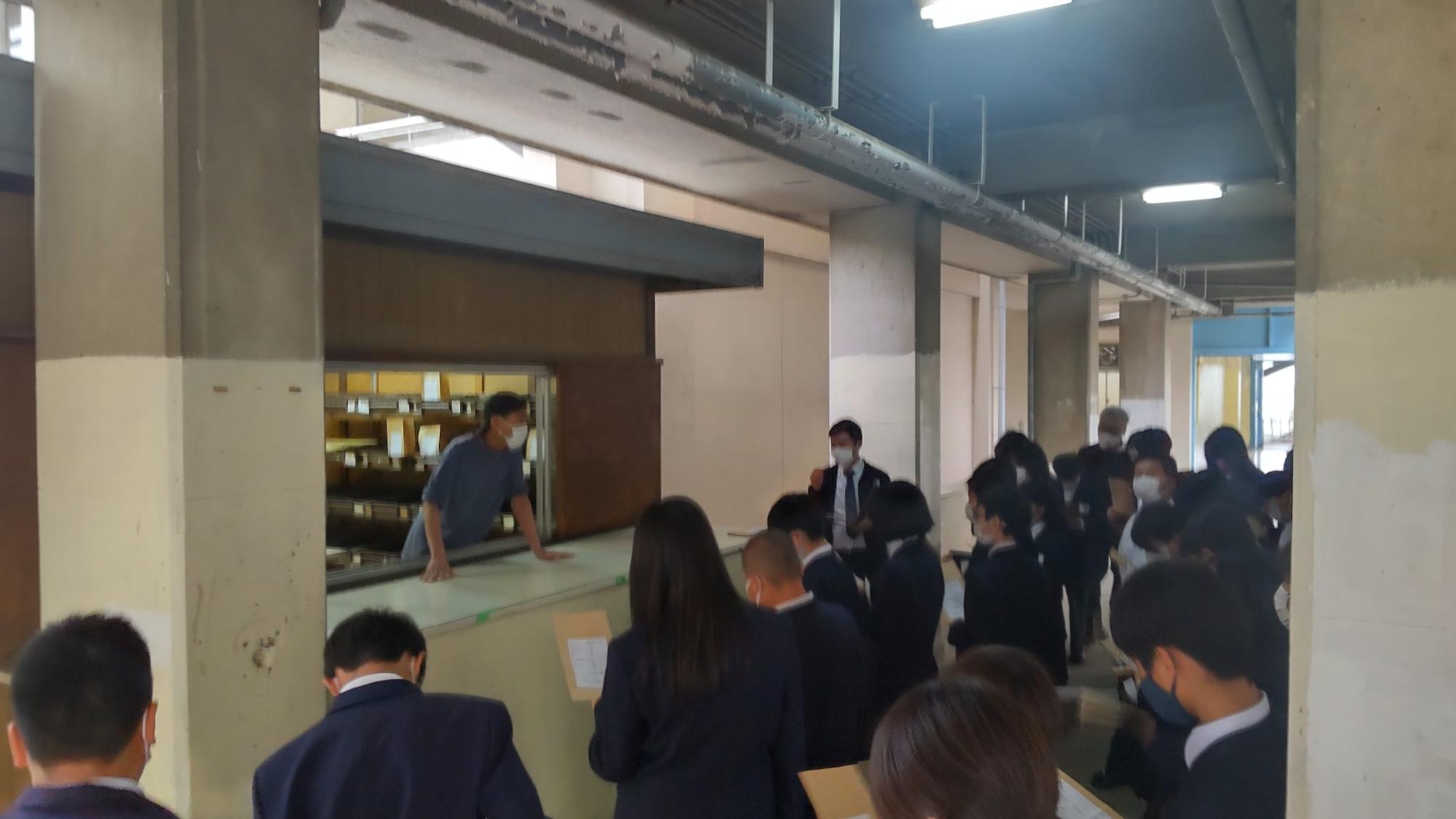 購買前の廊下に並んだ1年生の生徒たちがお店の男性からパンの購入の仕方の説明を聞いている様子の写真
