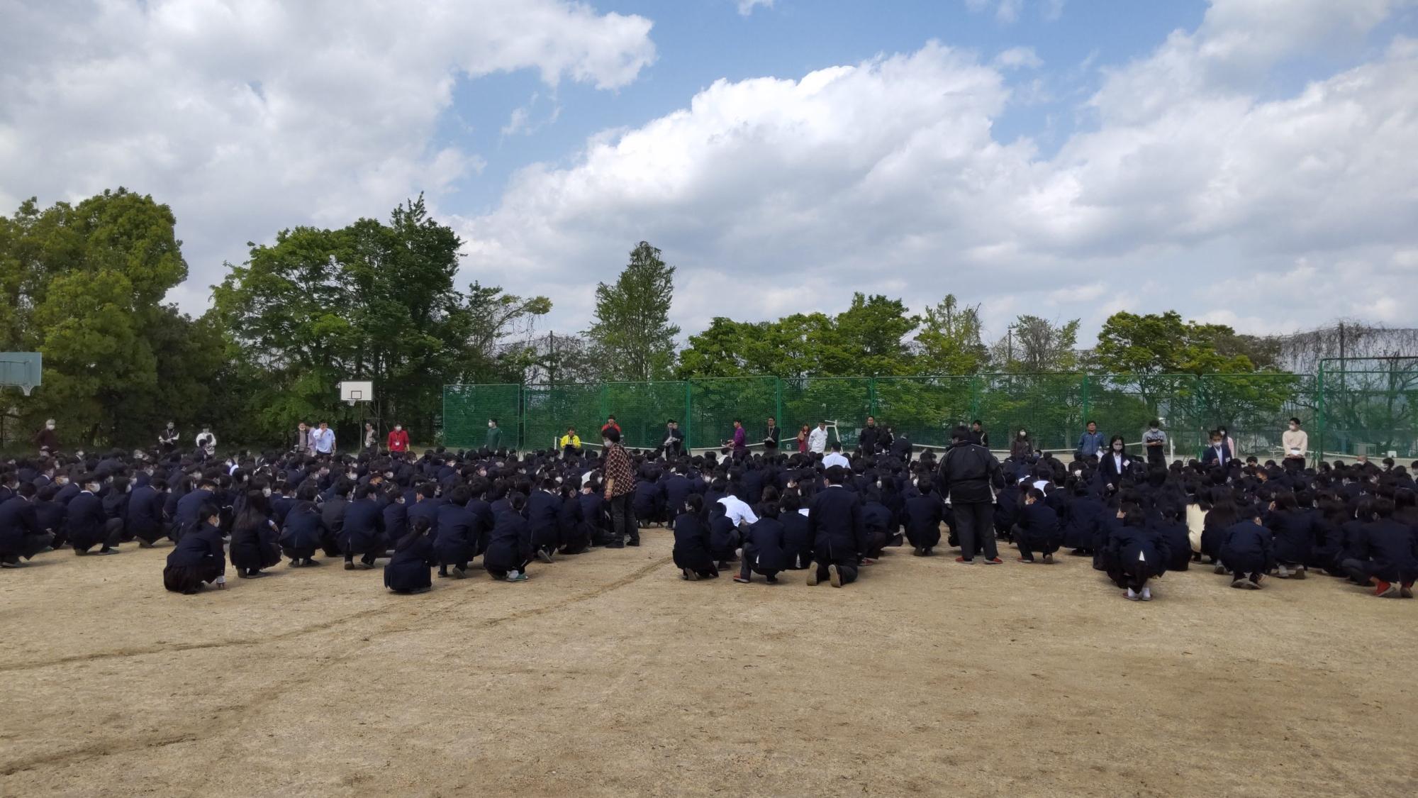 グランドの前方に教師が並び、制服姿の生徒たちがクラスごとに並んで座っている避難訓練後の集会の様子の写真