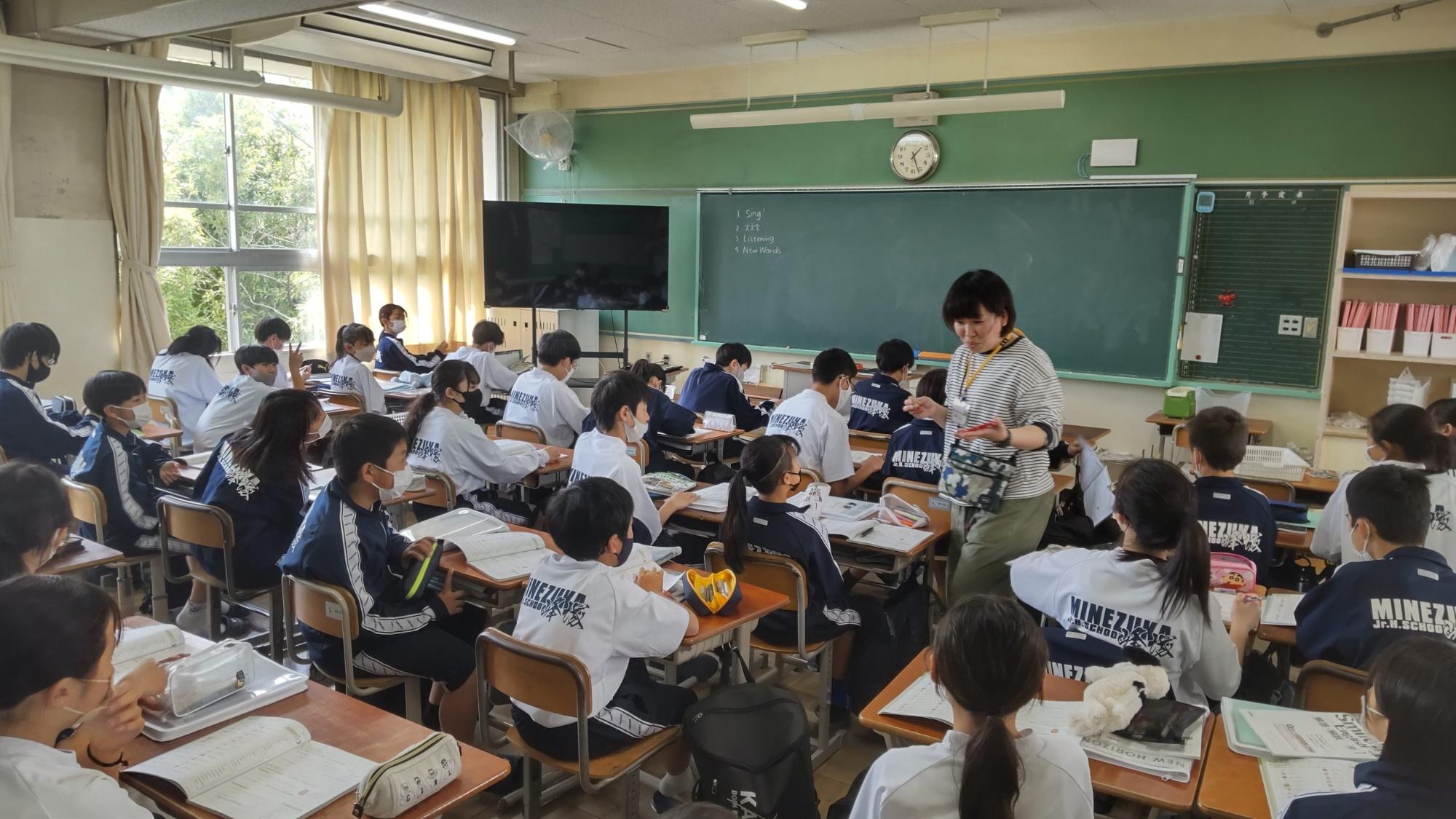 教室の自席に体操着姿の生徒たちが座り、女性教師が机の間を歩いている英語の授業の様子の写真