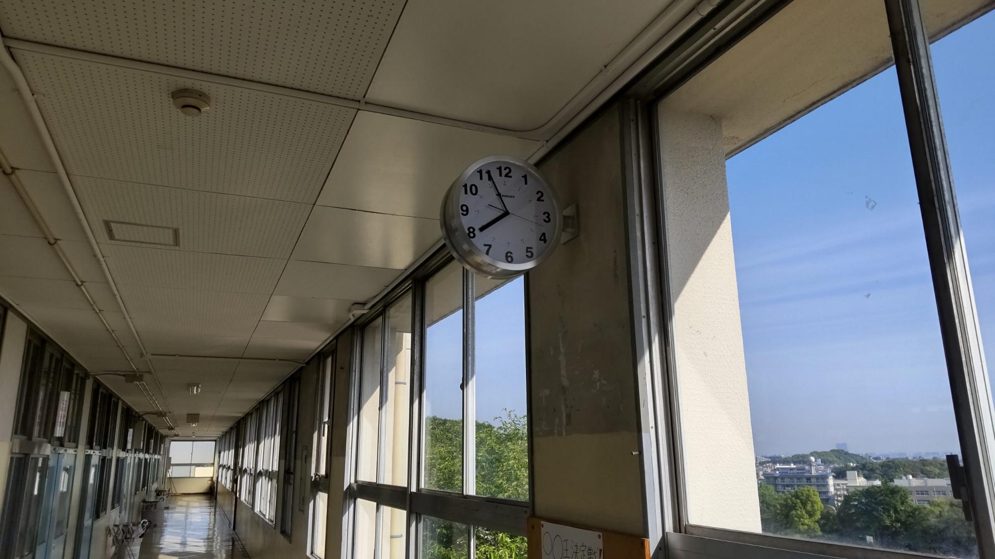 廊下の窓ガラス側の柱に設置された7時56分を指している寄贈された円形のスチール製の時計の写真