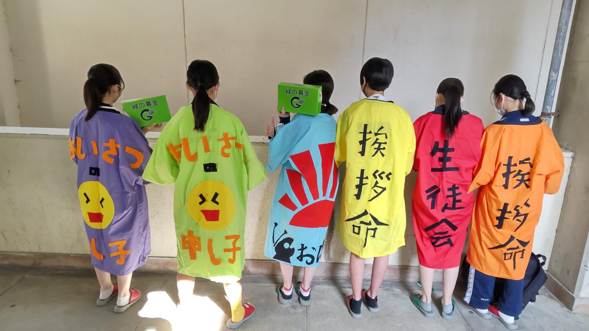 紫色や黄色などの法被の背中に「あいさつ申し子」や「挨拶命」と大きく書かれた生徒会の6名が横一列に並び、一番左と左から3番目に立つ生徒が緑の募金箱を持っている後ろ姿の写真