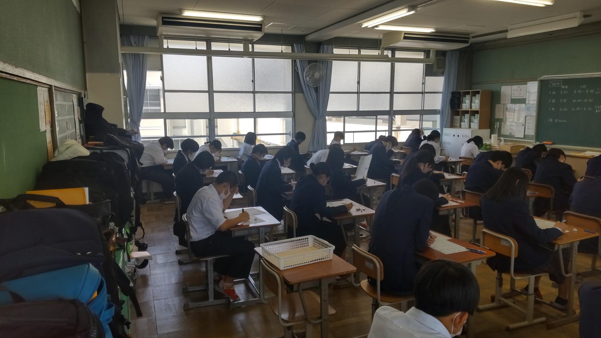 黒板にテストのスケジュールが書かれた教室の自席に座った生徒たちが答案用紙に向き合っているテストの様子の写真