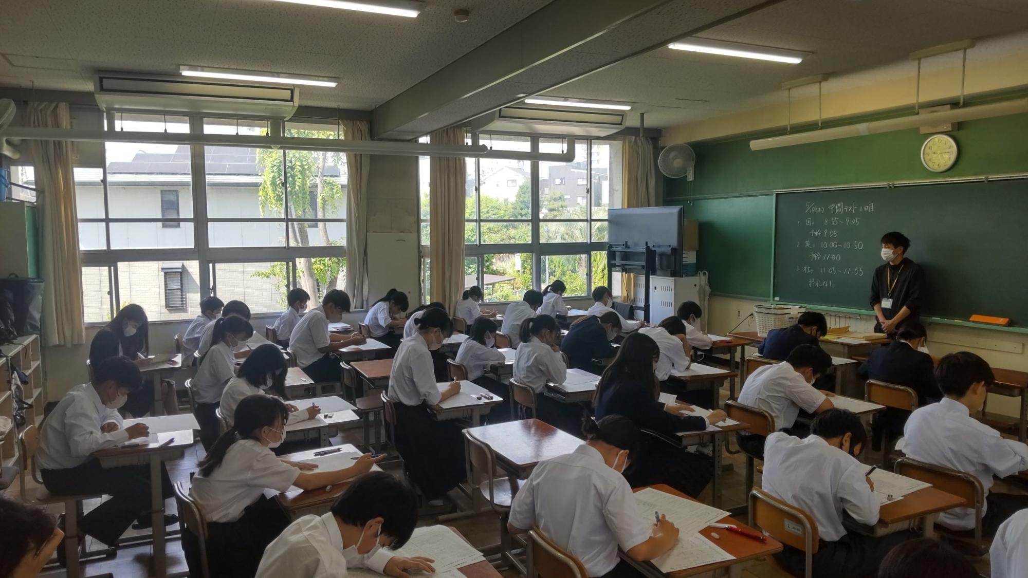 教室の黒板の前に男性教師が立ち、自席に座った生徒たちがテストを受けている様子の写真