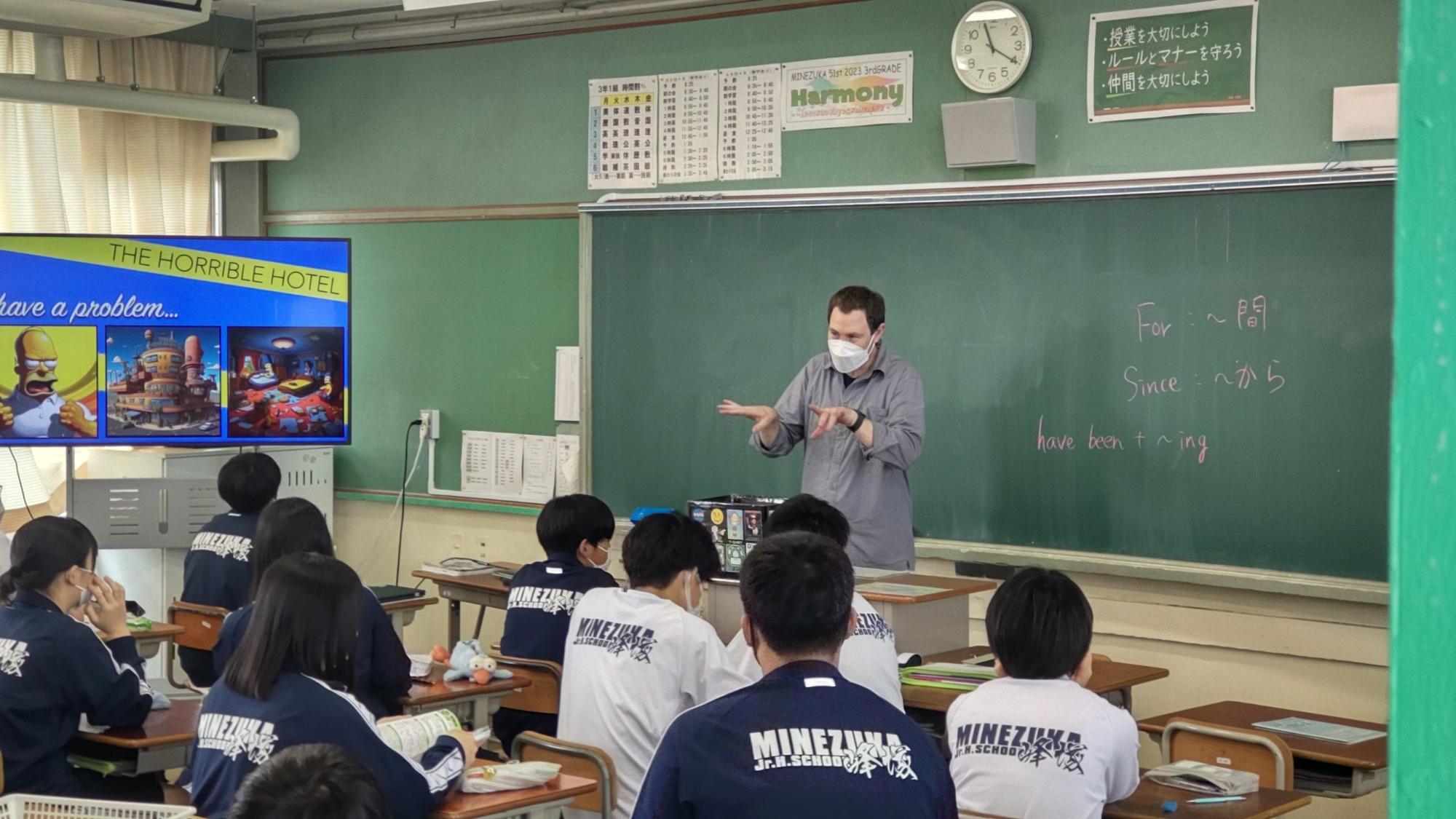 教室の左前方に設置されたモニターにアニメの映像が映し出され、自席に座った体操着姿の生徒たちが、黒板の前に立ち両手を前に出しているアンディー先生の話を聞いている授業の様子の写真