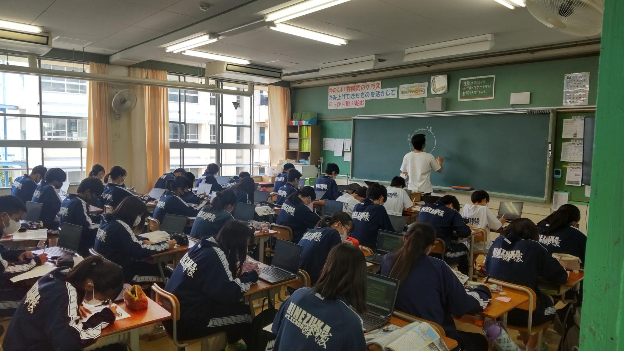 男性教師が白のチョークで黒板に円形を書き、自席に座っている生徒たちが机の上に教科書とタブレットを開き授業を受けている様子の写真