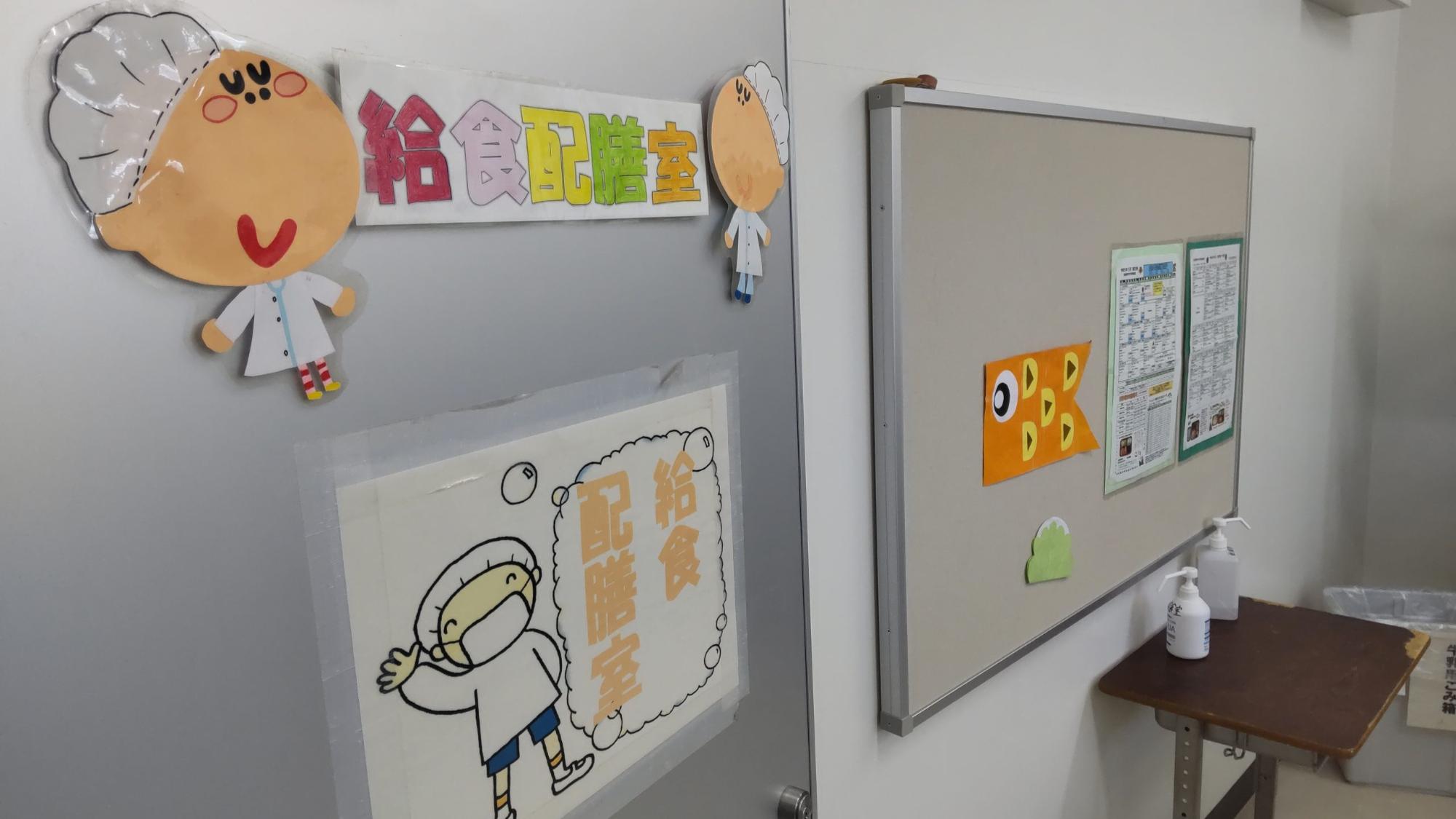 左側の扉に給食配膳室と書かれた手作りのイラストや飾りが貼られ、右側の掲示板に手作りのオレンジ色のこいのぼりと柏餅の飾りやメニュー表が貼られ、手前にゴミ箱と2つの消毒液が置かれた机が設置されている給食配膳室の写真