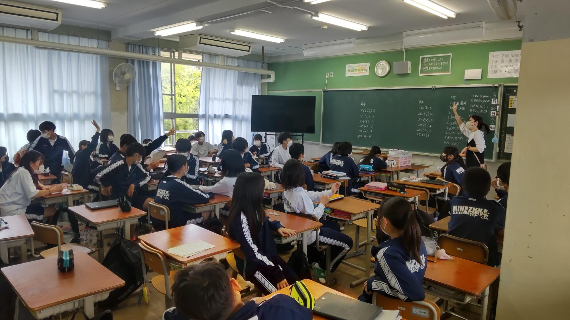 黒板に生徒の名前が書かれた教室前方に立つ女性教師が右手を挙げ、生徒たちが自席に座り窓側に座っている2名が手を挙げているクラスの取り組みの様子の写真