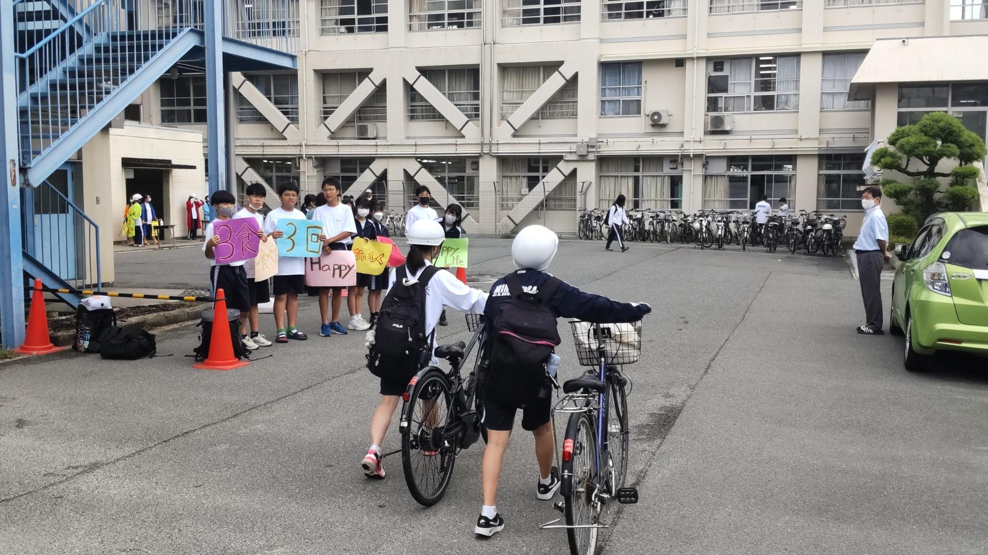 ヘルメットを被り敷地内を自転車を押して登校してくる2名の女子生徒に向かって保健委員会の1年生の7名が、3食食べて3回Happyなどの文字が書かれた色画用紙を持って呼びかけている様子の写真