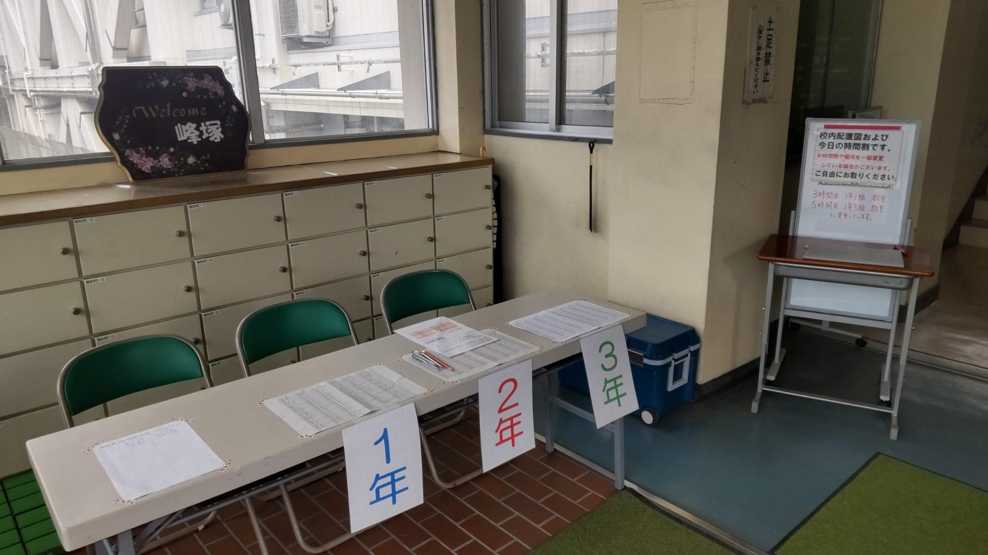 学校の入り口に3脚の椅子と長机が設置され、学年ごとに受付表が貼られている公開授業の受付の写真