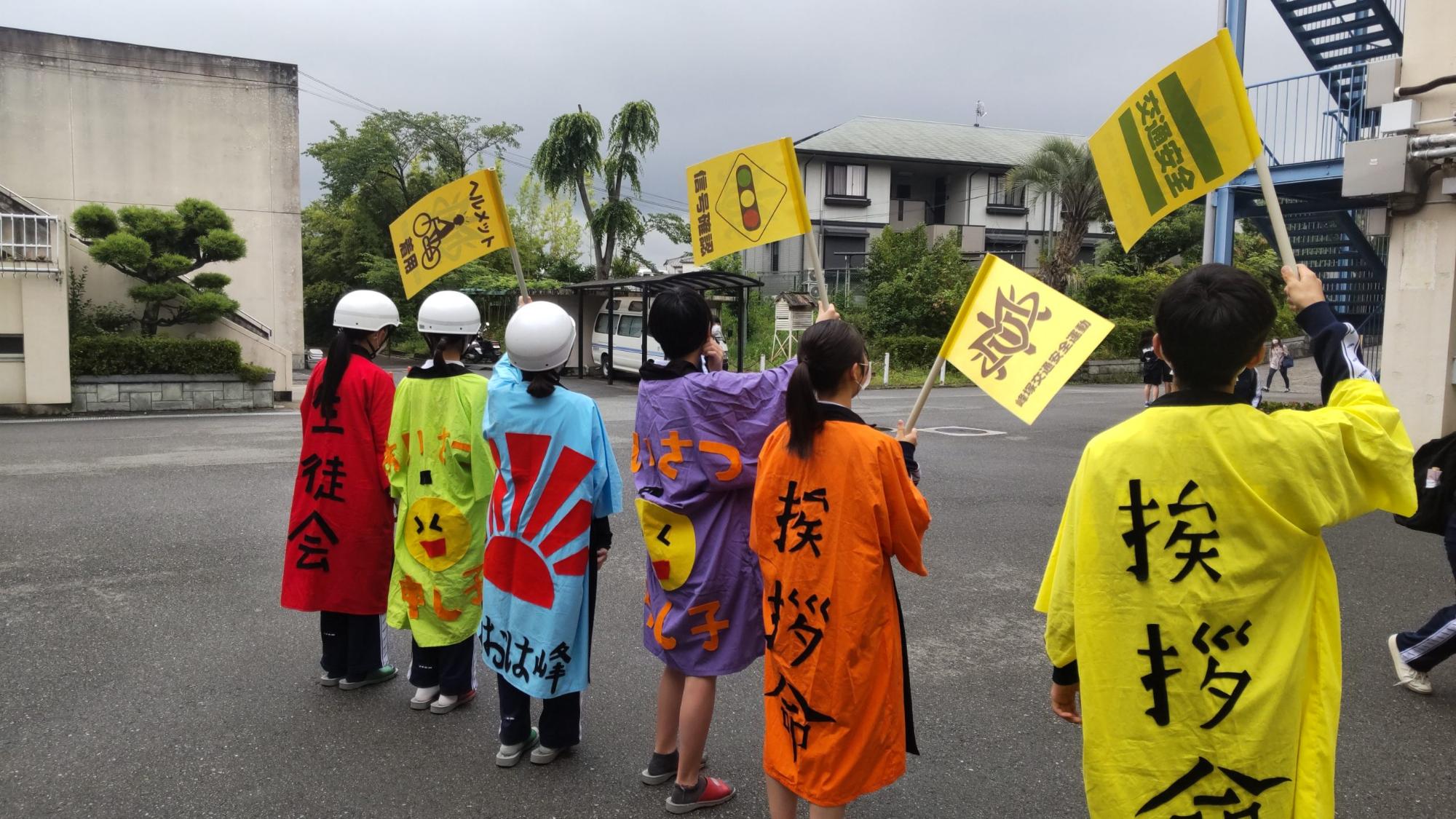 赤や黄色などの法被を着た6名の生徒会が横一列に並び、左側の3名はヘルメットを被り手作りの旗を振ってあいさつ運動を行っている様子の写真