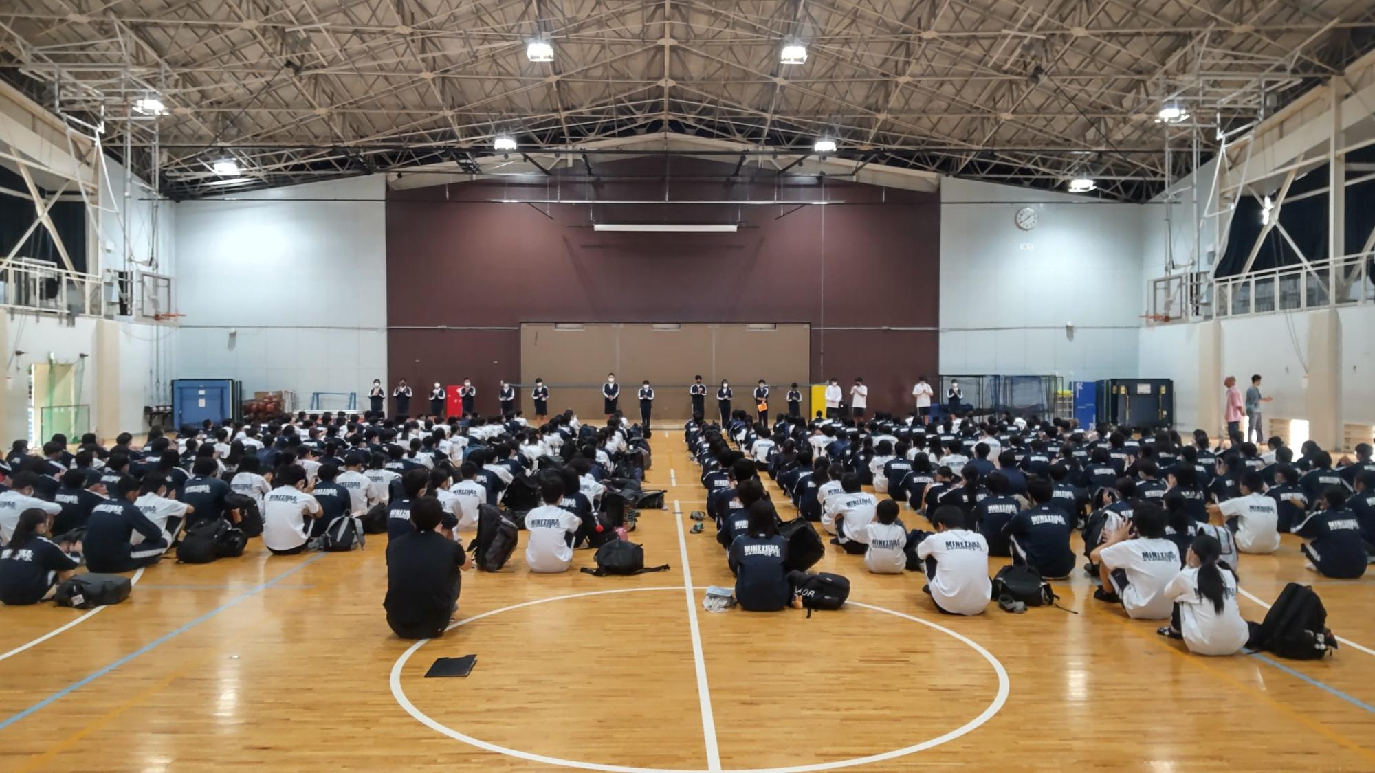体育館の前方に16名の生徒が横一列に並び、他の生徒たちが床に体育座りをして話を聞いている直前学年集会の様子の写真