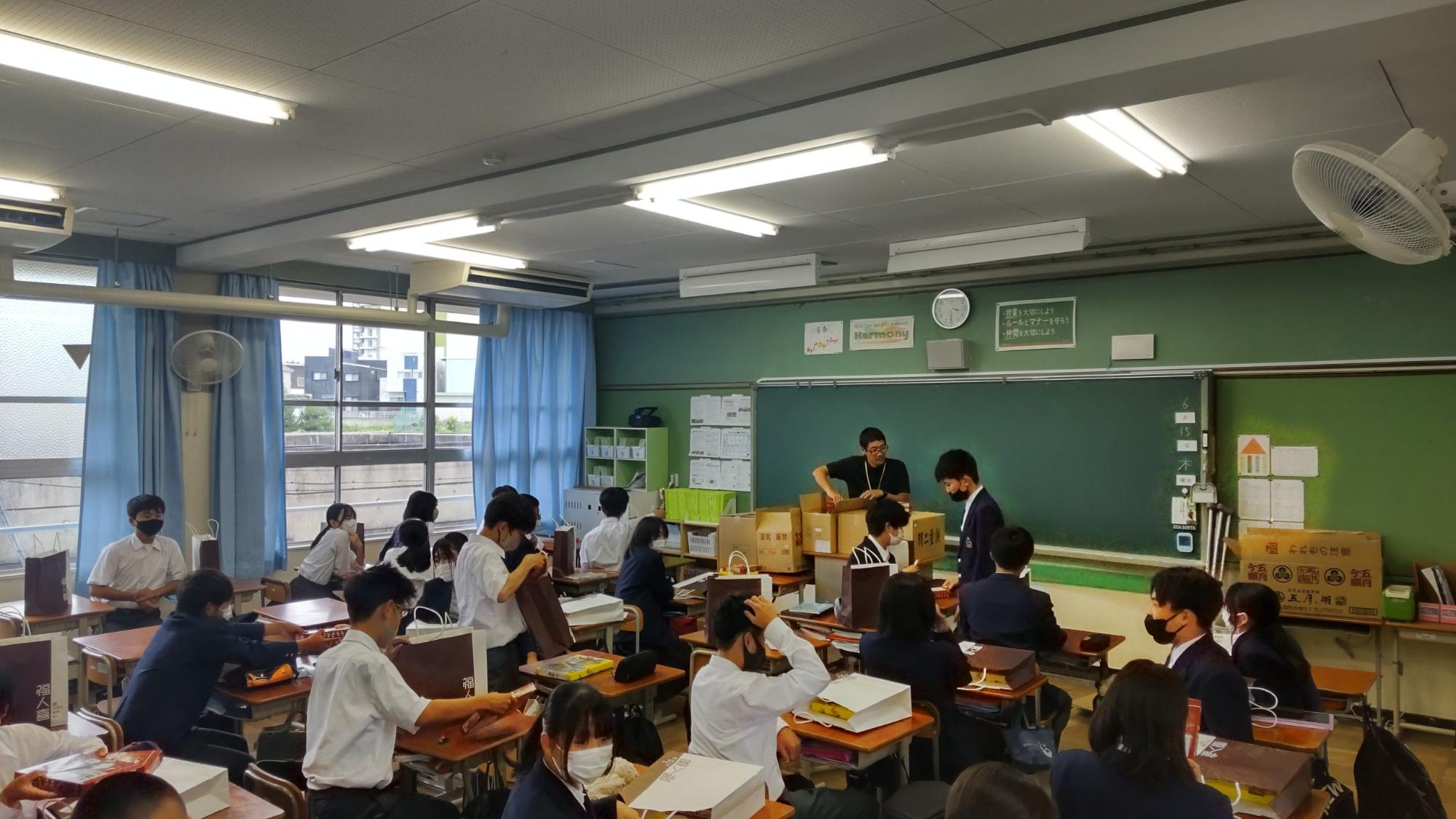 教室で男性教師が教壇の上に置かれた5箱の段ボールをあけ、自席に座っている生徒たちの机の上に配られたお土産が入った紙袋が置かれている写真