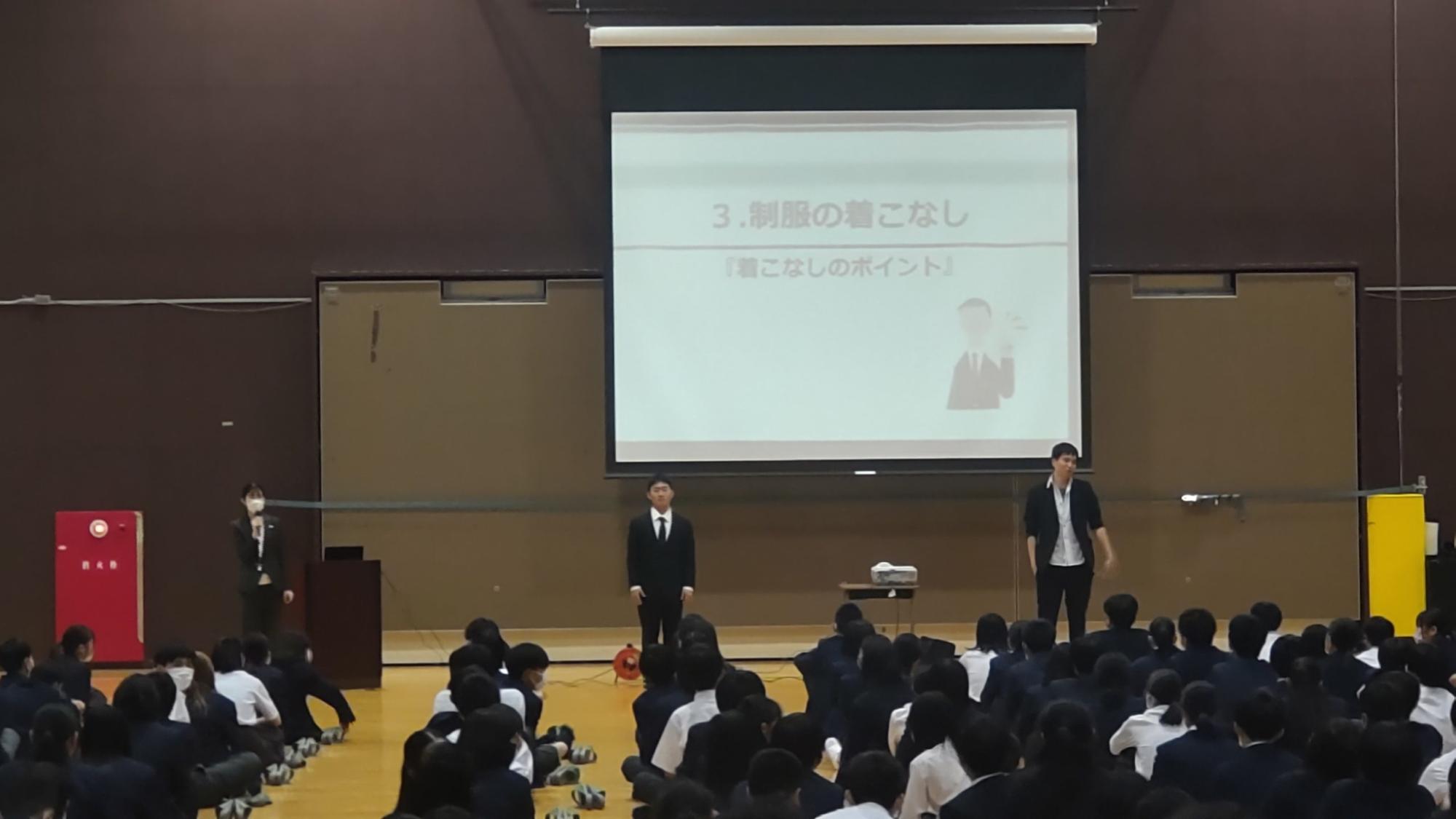 体育館の前方に「3.制服の着こなし「着こなしのポイント」」と書かれた資料が映し出された大きなスクリーンの前に2名の男子生徒が立ち、左側の男子生徒はネクタイを締めきちんと着こなしている一方、右側の男子生徒はジャケットやシャツの上のボタンやネクタイをはずし、ジャケットの袖をまくっている悪い着こなしのモデルを務め、左側の演台の前に立つ女性担当者の方の話を床に座った生徒たちが熱心に聞いている講演会の様子の写真