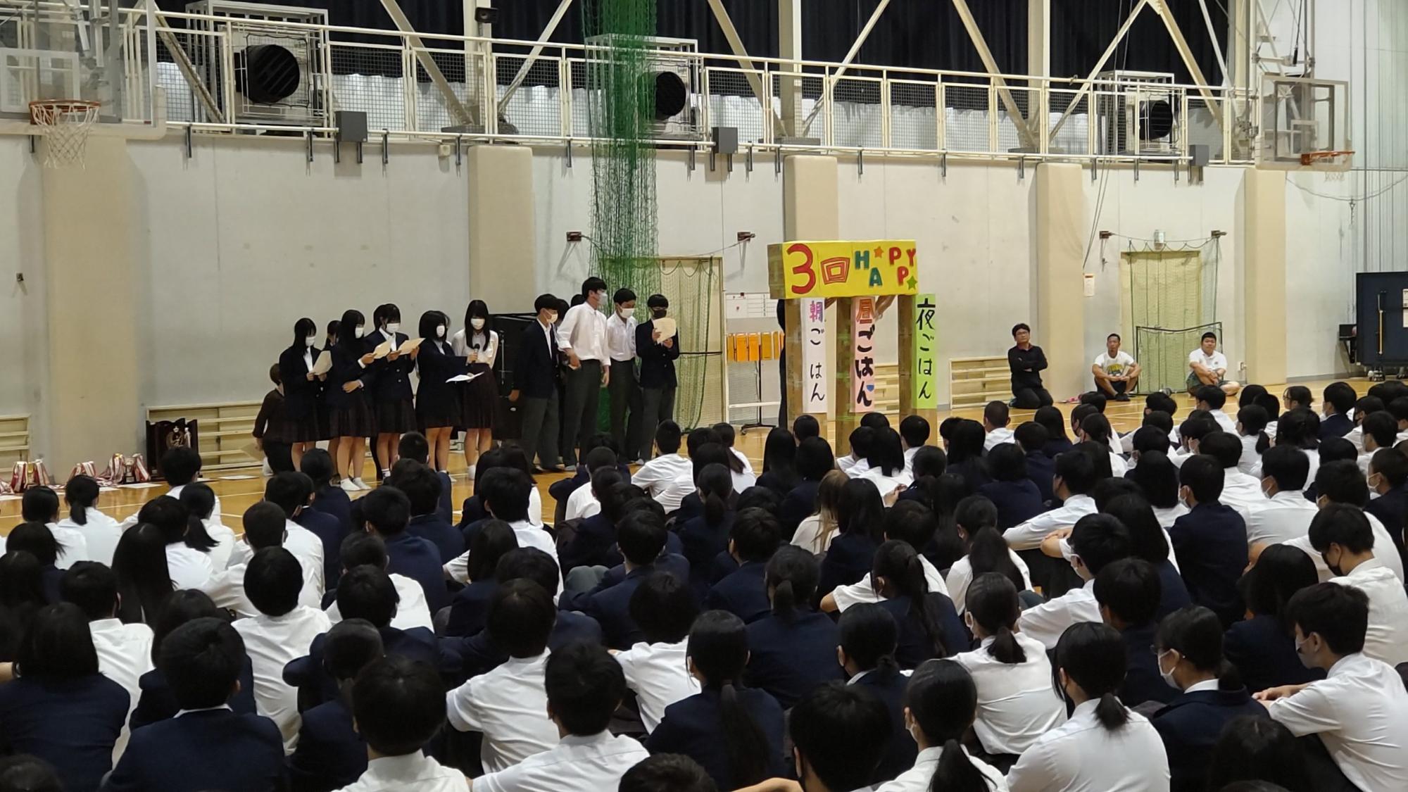 体育館の前方に「朝ごはん昼ごはん夜ごはん」と縦書きで書かれた手作りの柱が立ち、その上に大きく「3回HAPPY」と書かれた文字が置かれ、傍に立っている保健委員会の生徒たちが発表を行っている様子を他の生徒たちが床に座って聞いている写真