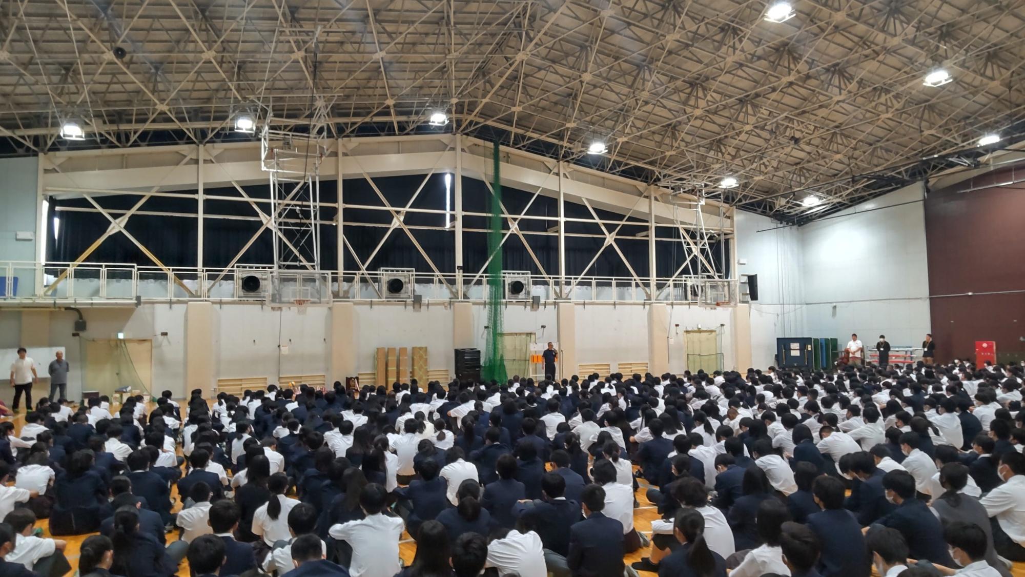 体育館の床に広がって座った生徒たちが、前方中央に立つ校長先生の話を聞いている後ろ姿の写真