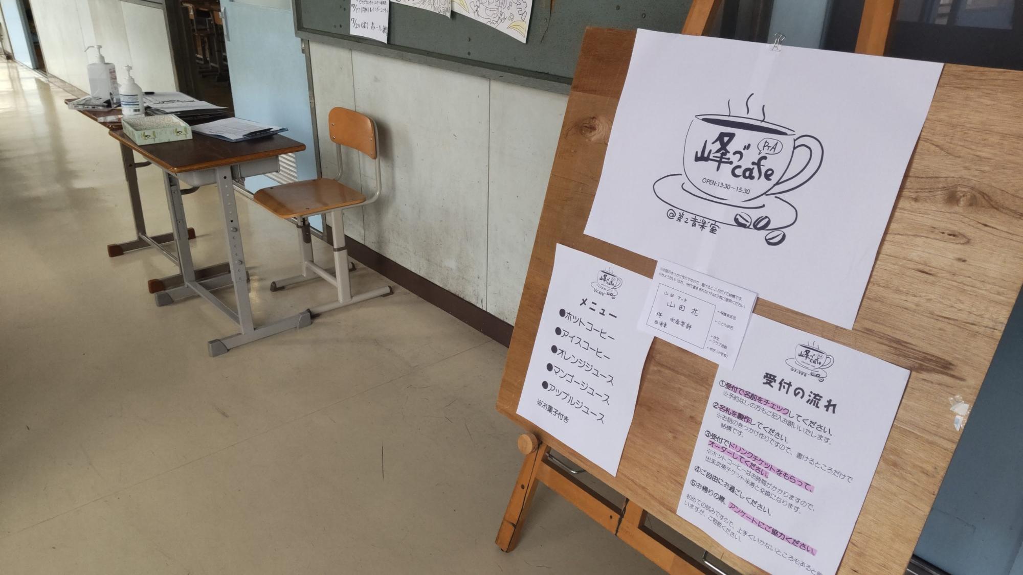 音楽室の会場の入り口の廊下に消毒液や受付表が置かれた2台の机が並び、右側に設置されたイーゼルに峰づcafeのデザイン画が描かれたイラストやメニュー、受付の流れが書かれた用紙が貼られている手作り看板の写真