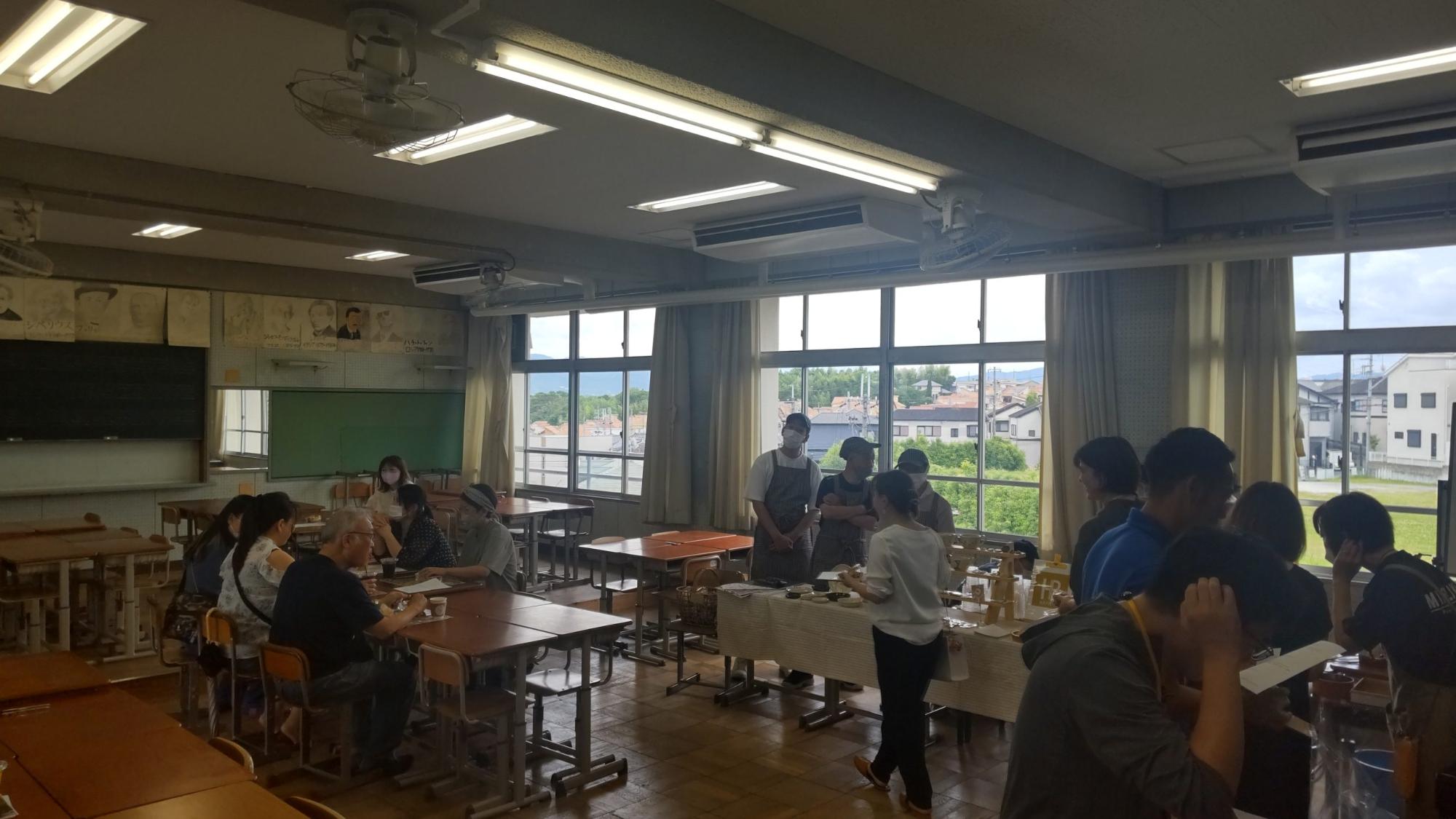 音楽室の窓側に机が並べられテーブルクロスが敷かれたブースにカフェに来店したお客さんが並び、机をつけてグループごとに座っているお客さんがスイーツや飲み物を頂いている峰づcafeの様子の写真