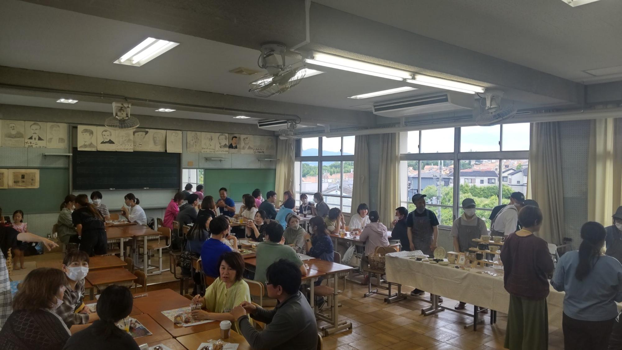音楽室の窓側に机が並べられテーブルクロスが敷かれた上にコーヒーカップなどが並んだブースに「ハピバール」のスタッフの方々が立って接客を行い、机をつけてグループごとに座っているお客さんがスイーツや飲み物を頂いている満席状態の峰づcafeの様子の写真