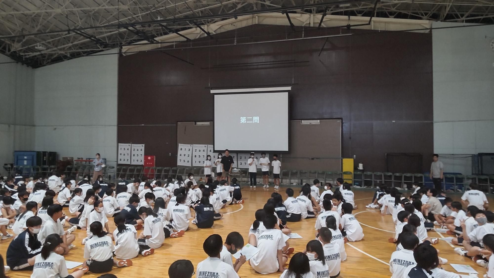 体育館の前方に「第二問」と表示された大きなスクリーンの下に男性教師tと5名の生徒が立ち、他の生徒たちが床に座っている学年レクの様子の写真