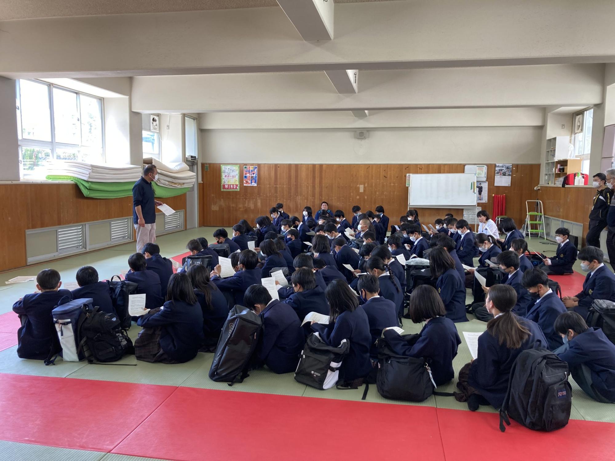 柔道場の前方に男性教師が立ち、床に座った1年生の生徒たちが話を聞いている自転車講習会の様子の写真
