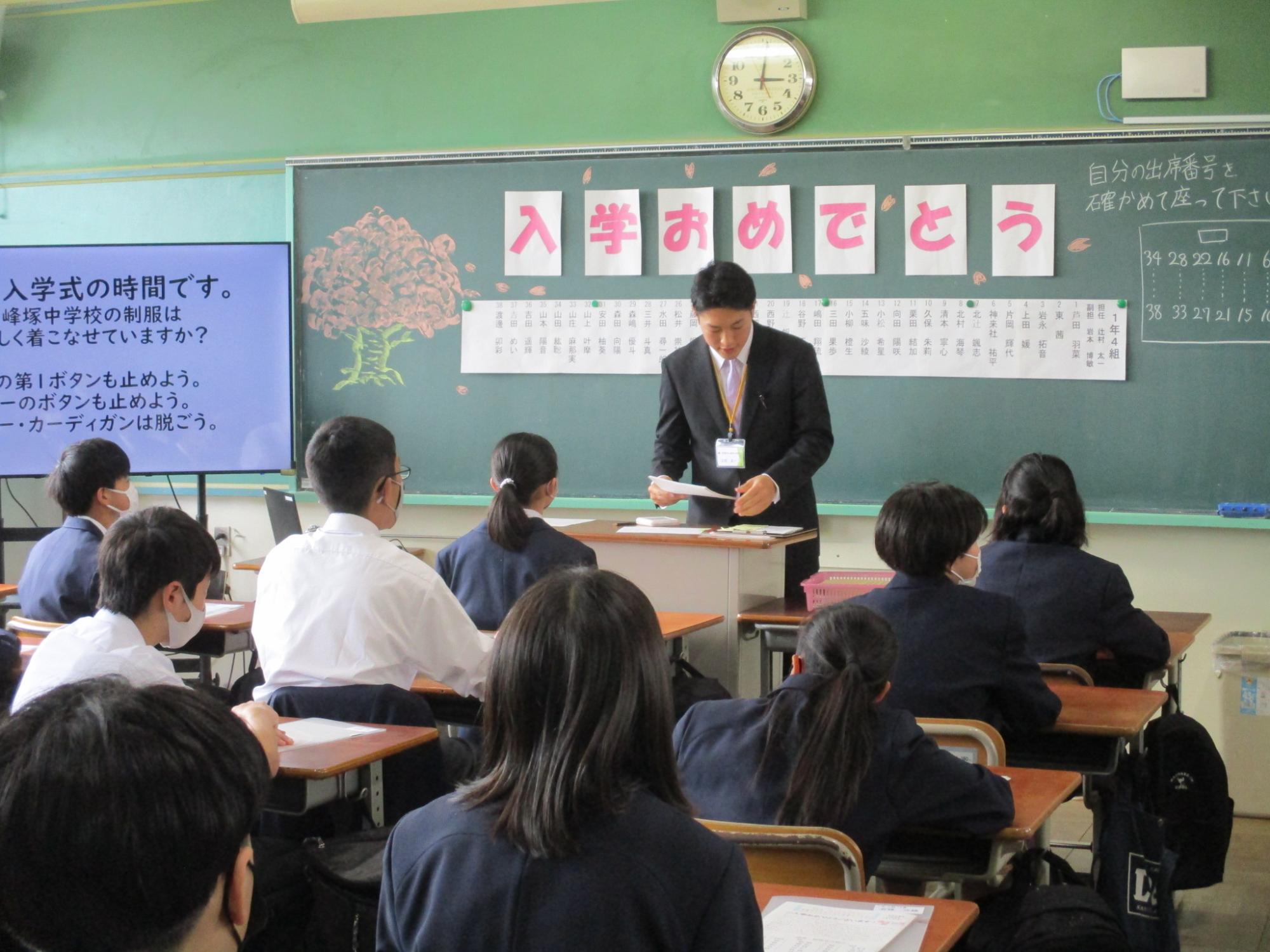 ピンク色の文字で一文字ずつ「入学おめでとう」と書かれた用紙とクラス名や担任と副担任、38名の生徒の名前が書かれた紙が貼られた黒板の前の教壇にスーツ姿の男性教師が立ち、自席に座った生徒たちが先生の話を聞いている教室の様子の写真
