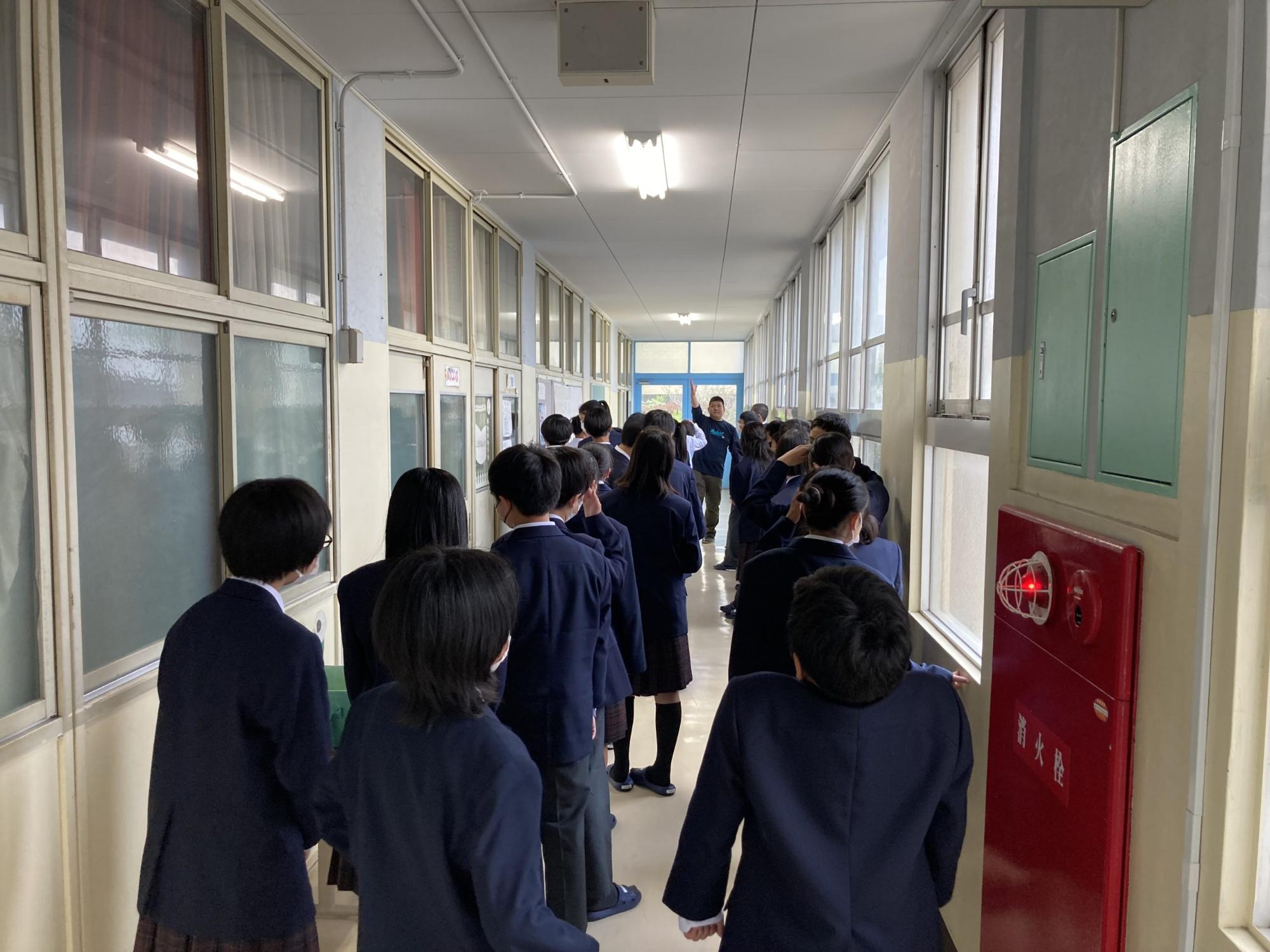 廊下に2列に並んだ1年生の生徒たちが、先頭で右手を上げて立つ男性教師の話を聞いている校舎探検の様子の写真