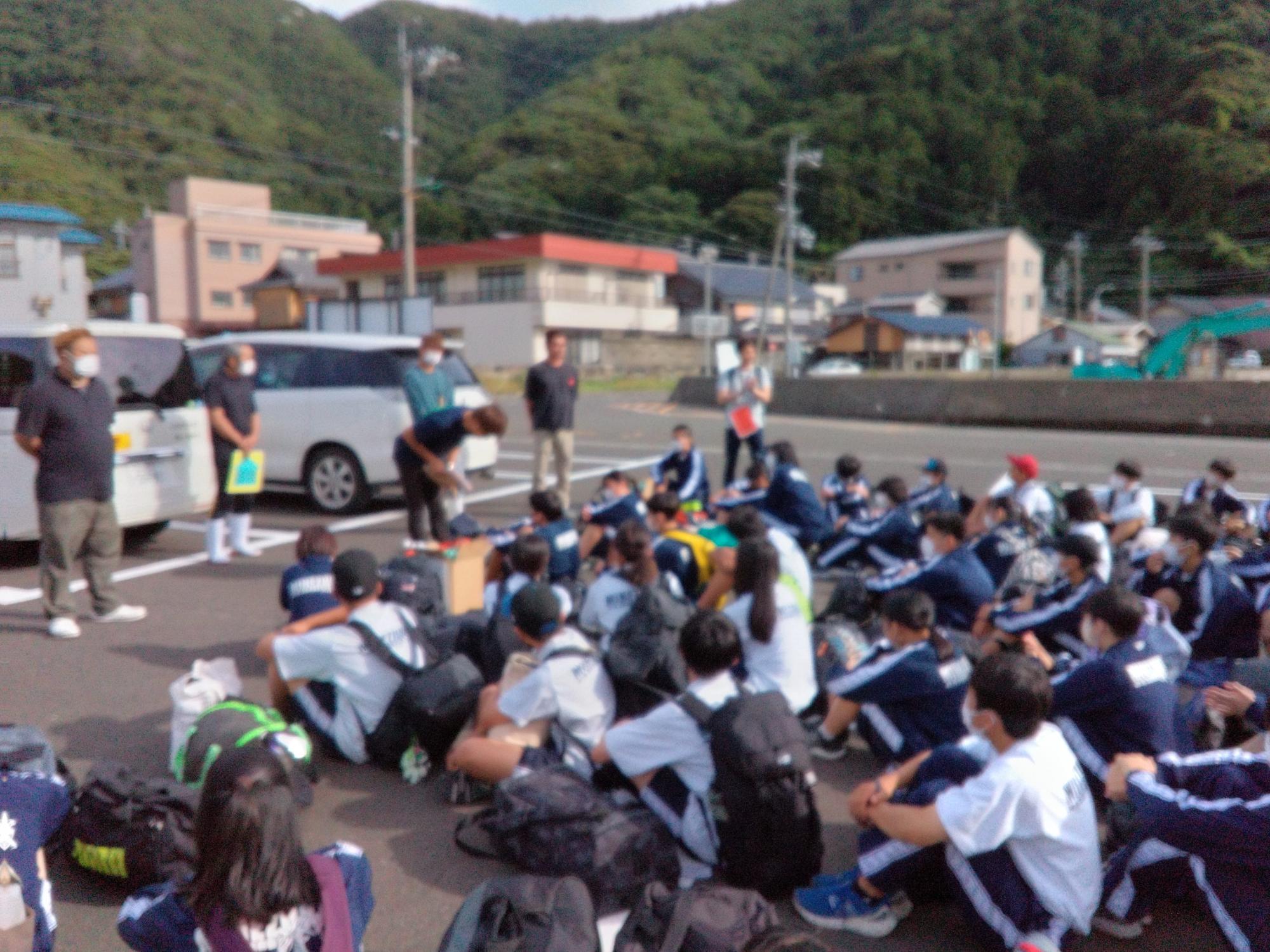 宿の駐車場に到着した生徒たちが体育座りをして先生方の話を聞いている様子の写真