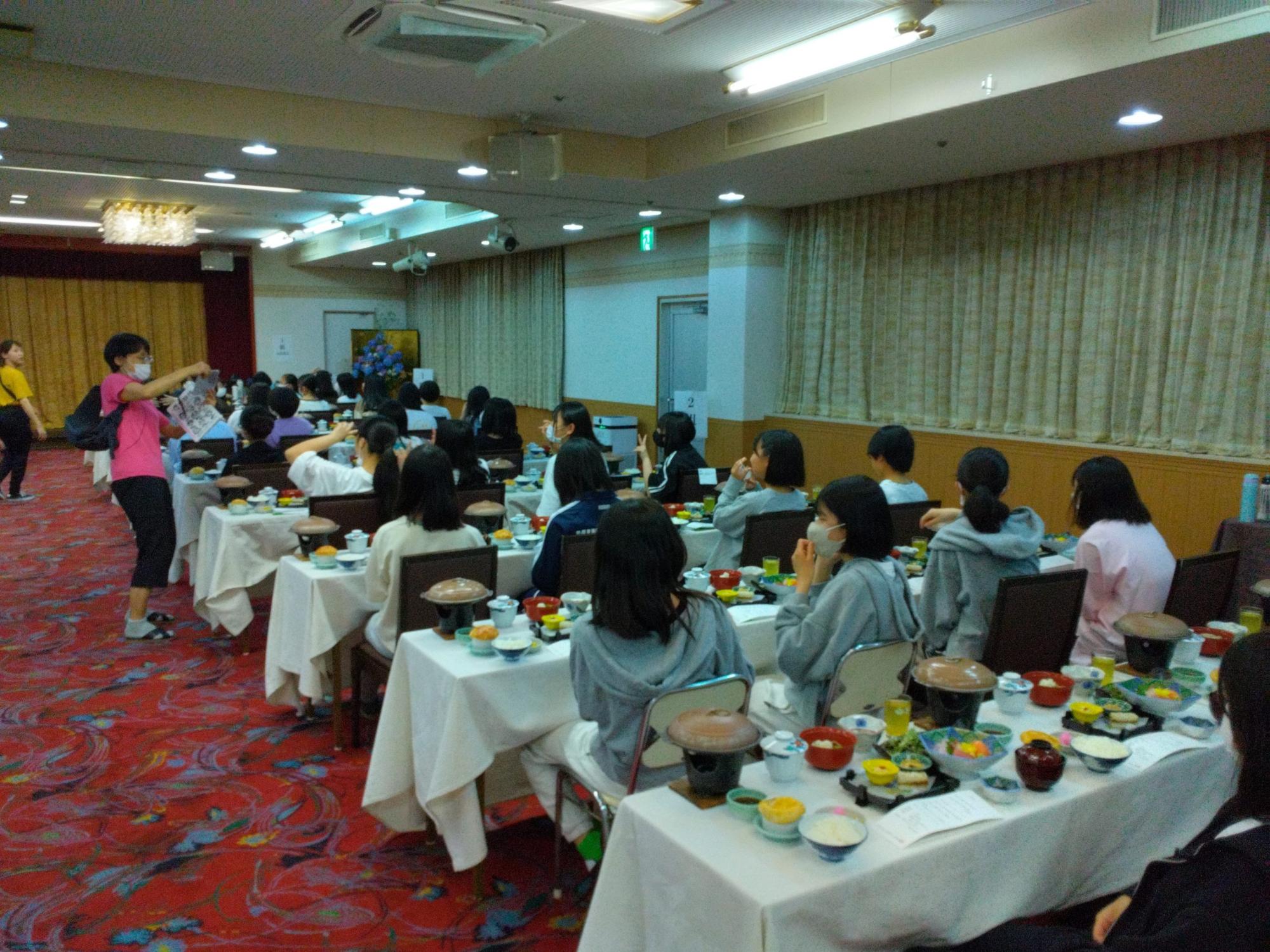 4名掛けのテーブルの上に食事が並び、着席した女子生徒たちが左側に立っている女性教師の話を聞いている後ろ姿の写真