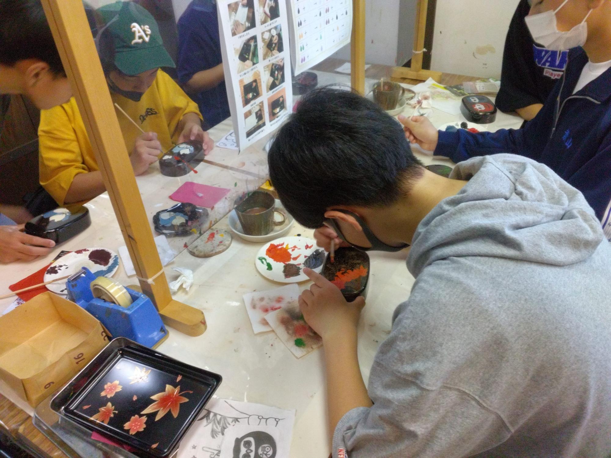 男子生徒が絵が描かれたお弁当箱の蓋に塗料を塗っている伝統工芸の蒔絵体験を行っている様子の写真