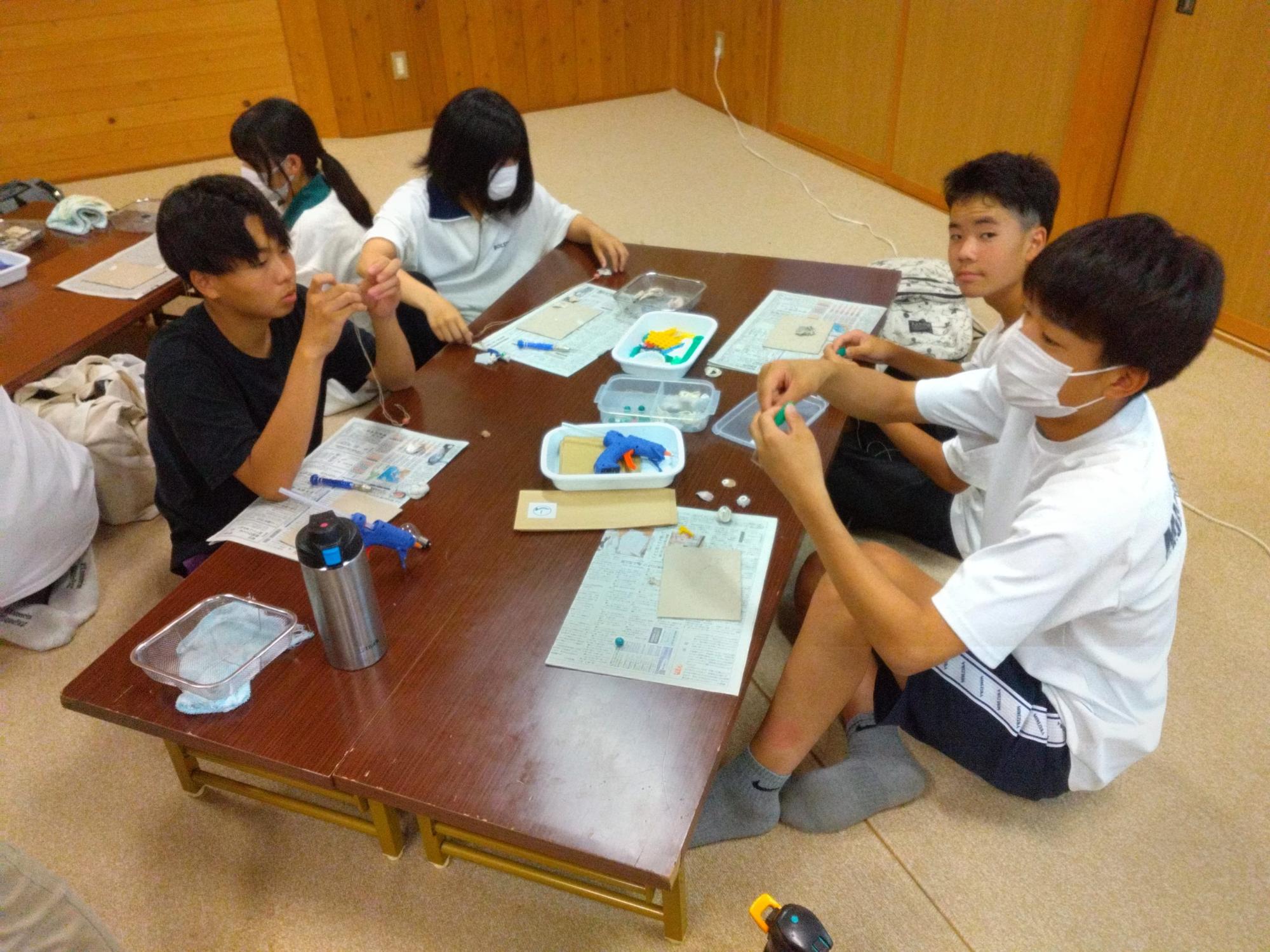 2台の長机をつけて4名ずつ座った生徒たちが、グルーガンを使用し細かい作業を行っている様子の写真