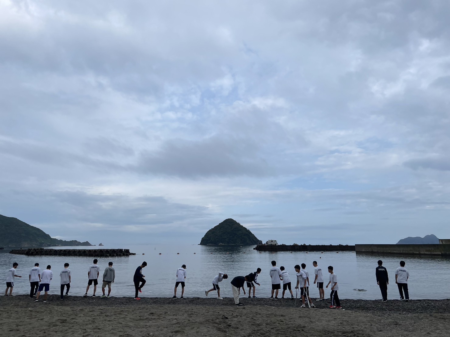 湖岸に横一列に並んだ男子生徒が、山形の小島が浮かぶ湖に向かって石を投げている後ろ姿の写真