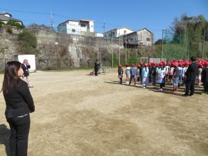 黒いスーツ姿の先生が手前に立っており、奥には赤い帽子をかぶった児童の皆さんが整列している赴任式・始業式の様子を写した写真