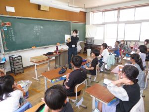 教材を手に持ち、指さしして説明している先生、子どもたちは自席につき先生の話を聞いている授業風景を写した写真