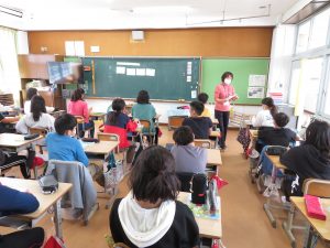 教室で黒板には教材が掲示され、自席について授業に参加している子どもたちと、教材を手に持ち授業をしている先生が写っている教室の様子を写した写真