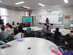丸いテーブルのある教室に子どもたちがグループに分かれて座っており、前方には映像が映し出されたモニターがあり、英語教員が子どもたちの前に立ち授業が行われている様子の写真