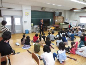 リコーダーの達人の男性が大きなリコーダーを演奏しており、3年生の子どもたちが演奏者の前に座って興味深く演奏を聞いているリコーダー講習会の様子の写真
