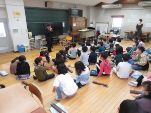 リコーダーの達人の男性が手のひらに収まる小さなリコーダーを演奏しており、3年生の子どもたちが座って演奏を聞いているリコーダー講習会の様子の写真