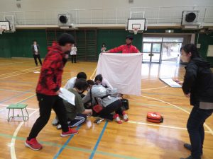 体育館で白い大きな布を広げて立つ先生、白い布の前に4名の先生が集まり、事故対応の訓練を行っており、近くで2名の先生が資料を手に持ち訓練の様子を確認している食物アレルギー研修の様子の写真