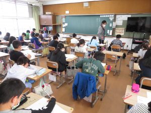 一人の子どもの様子を見守っている先生と、自席に座り集中して問題に取り組んでいる子どもたちの様子を写した写真