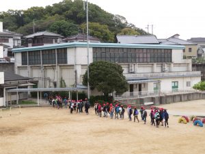 赤い帽子をかぶり、リュックを背負った子どもたちが列に並んで歩いており、運動場から校外活動に出発している様子を後方から写した写真