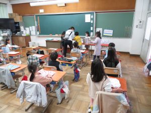 机の上にランチマットを広げ、自席に座ってまっている子どもたちと、4名の子どもたちと先生が黒板前の机に集まっては給食の配膳の手伝いをしてる給食の様子の写真