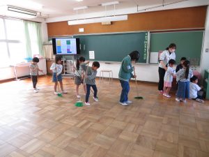 教室で、6年生の児童の後ろに4名の1年生が並んで、ほうきで床をはいて掃除をしている様子の写真