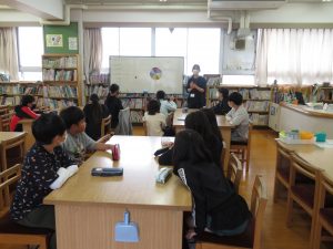 沢山の本が並んでいる図書室で、委員会の子どもたちがグループに分かれて席についており、ホワイトボードをの横で説明している先生の話を聞いている委員会活動の様子の写真